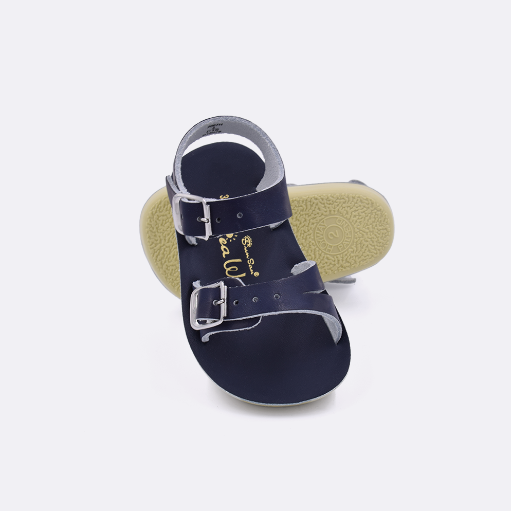 Two baby sized 2000 Sea Wee style sandals with navy straps and navy insoles.  One standing with the sole facing the camera. The second is laying diagonally over the top left edge of the sole.