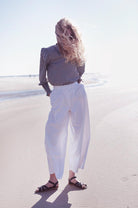An adult with blonde hair is standing on the beach with a checkered black and white shirt tucked into white pants. Their hands are in their pockets with their feet diagonally facing each direction wearing our Brown Salt Water Originals.
