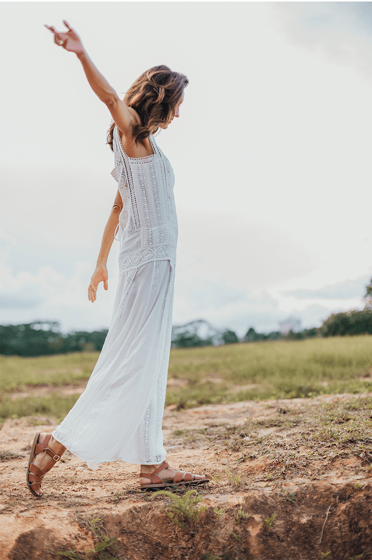 An adult with long brown hair walking on a dirt and grass terrain. They are wearing a white dress and our tan salt water original sandals while swinging their right arm in the air.