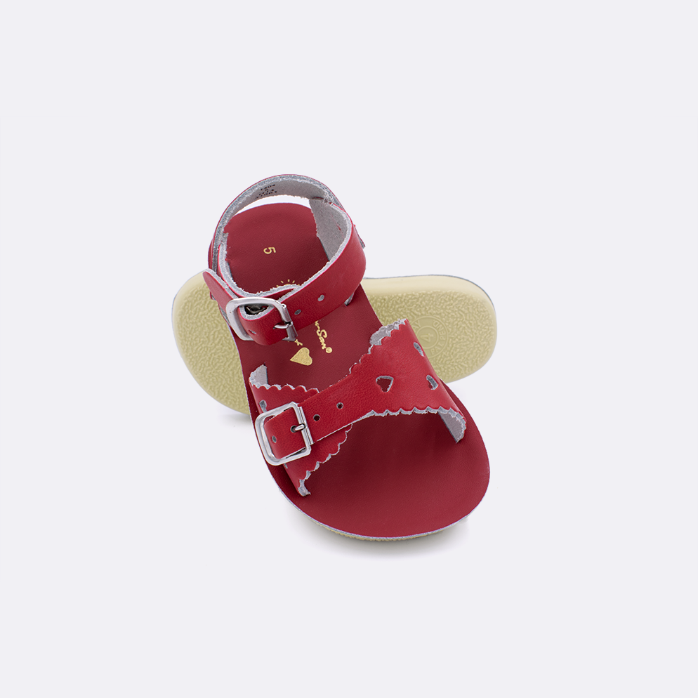 Two toddler sized 1400 Sweetheart style sandals with red straps and red insoles.  One standing with the sole facing the camera. The second is laying diagonally over the top left edge of the sole.