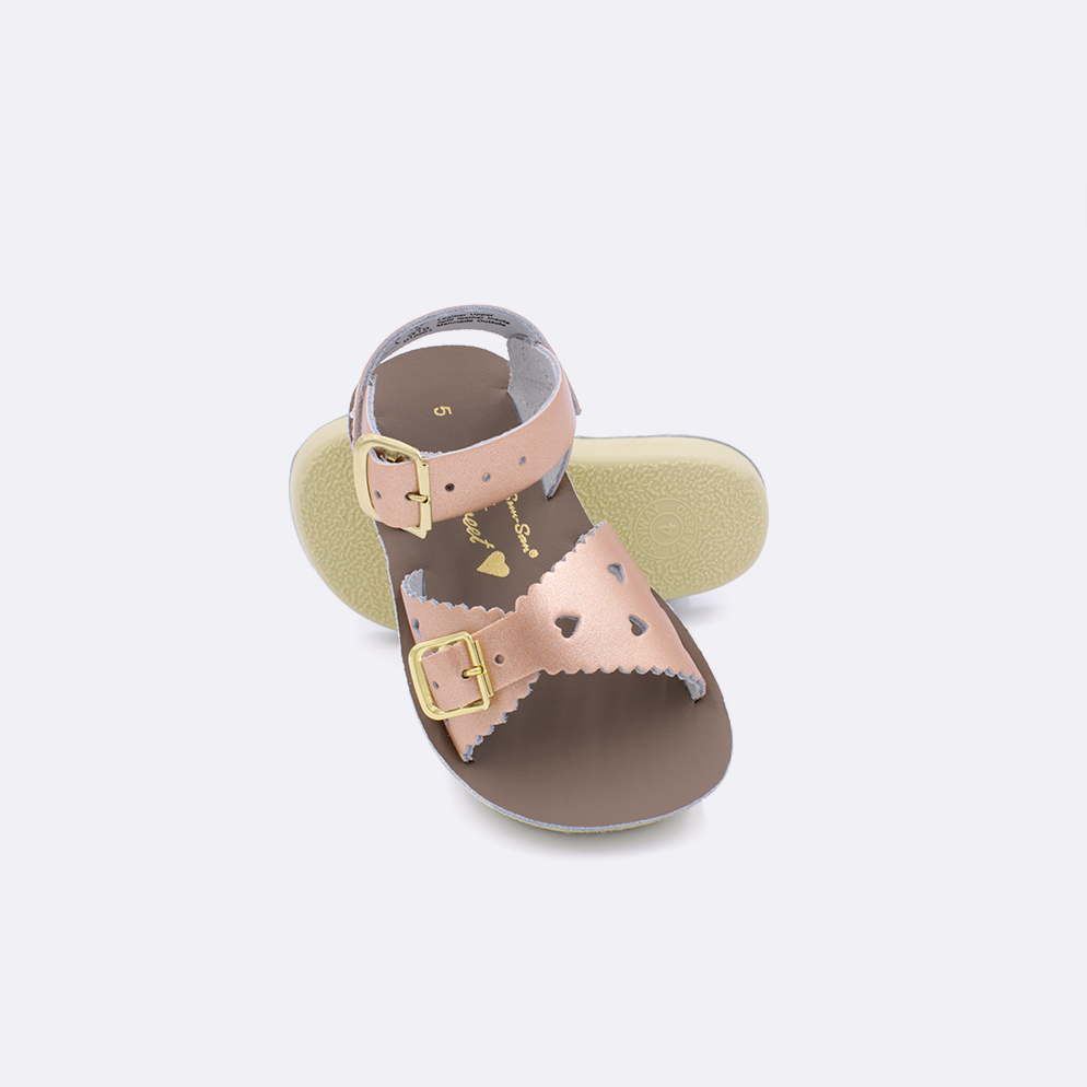Two toddler sized 1400 Sweetheart style sandals with rose gold straps and beige insoles.  One standing with the sole facing the camera. The second is laying diagonally over the top left edge of the sole.
