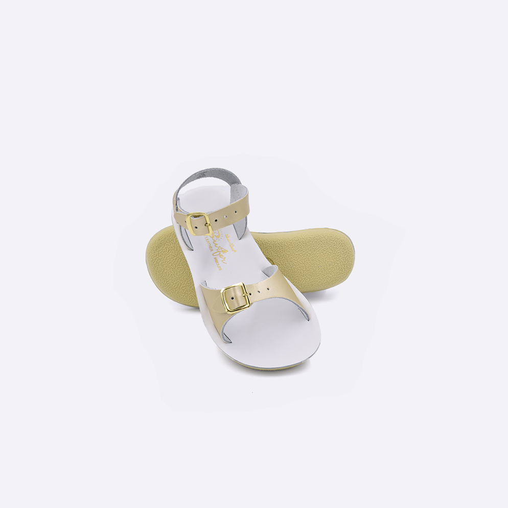 Two toddler sized 1700 Surfer style sandals with gold straps and white insoles.  One standing with the sole facing the camera. The second is laying diagonally over the top left edge of the sole.