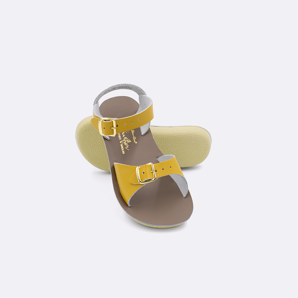 Two toddler sized 1700 Surfer style sandals with mustard straps and beige insoles.  One standing with the sole facing the camera. The second is laying diagonally over the top left edge of the sole.