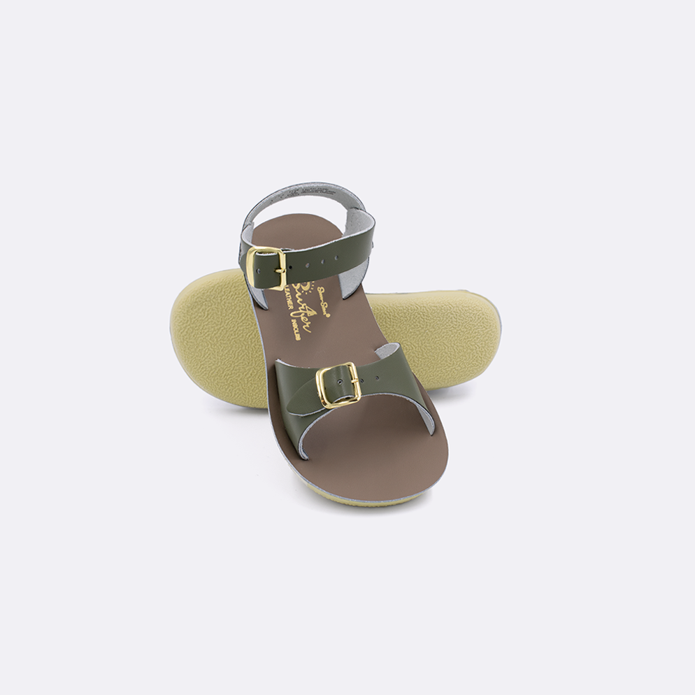 Two toddler sized 1700 Surfer style sandals with olive straps and beige insoles.  One standing with the sole facing the camera. The second is laying diagonally over the top left edge of the sole.