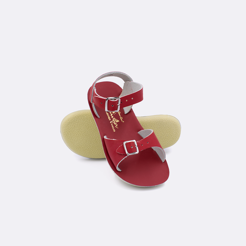Two toddler sized 1700 Surfer style sandals with red straps and red insoles.  One standing with the sole facing the camera. The second is laying diagonally over the top left edge of the sole.