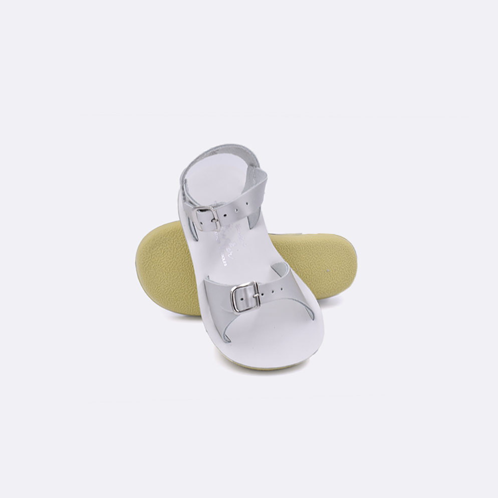 Two toddler sized 1700 Surfer style sandals with silver straps and white insoles.  One standing with the sole facing the camera. The second is laying diagonally over the top left edge of the sole.