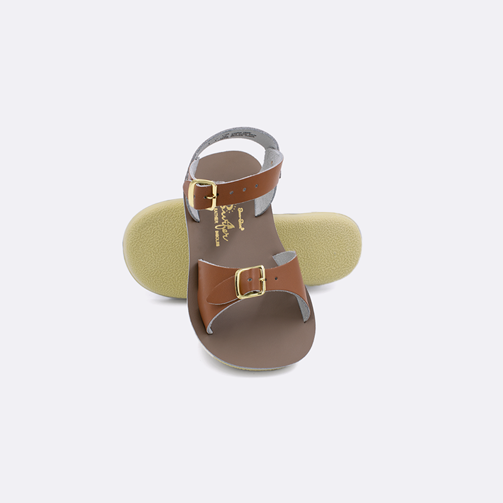 Two toddler sized 1700 Surfer style sandals with tan straps and beige insoles.  One standing with the sole facing the camera. The second is laying diagonally over the top left edge of the sole.