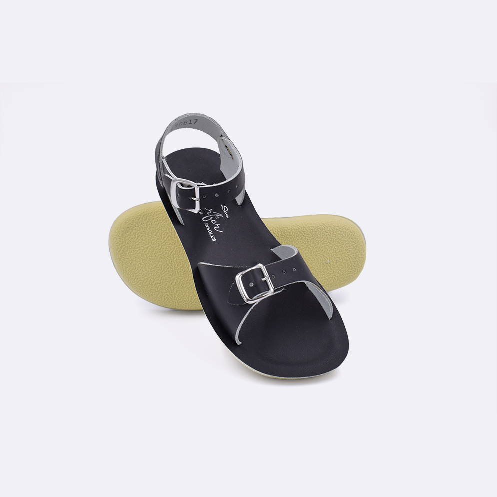 Two little kid sized 1700 Surfer style sandals with black straps and black insoles.  One standing with the sole facing the camera. The second is laying diagonally over the top left edge of the sole.