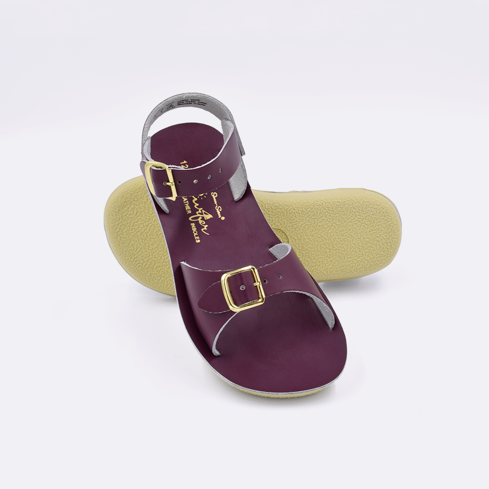 Two little kid sized 1700 Surfer style sandals with claret straps and claret insoles.  One standing with the sole facing the camera. The second is laying diagonally over the top left edge of the sole.