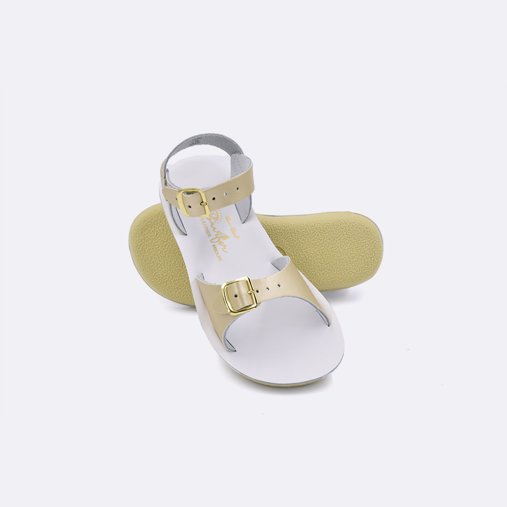 Two little kid sized 1700 Surfer style sandals with gold straps and white insoles.  One standing with the sole facing the camera. The second is laying diagonally over the top left edge of the sole.