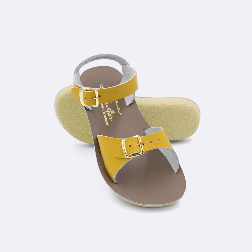 Two little kid sized 1700 Surfer style sandals with mustard straps and beige insoles.  One standing with the sole facing the camera. The second is laying diagonally over the top left edge of the sole.