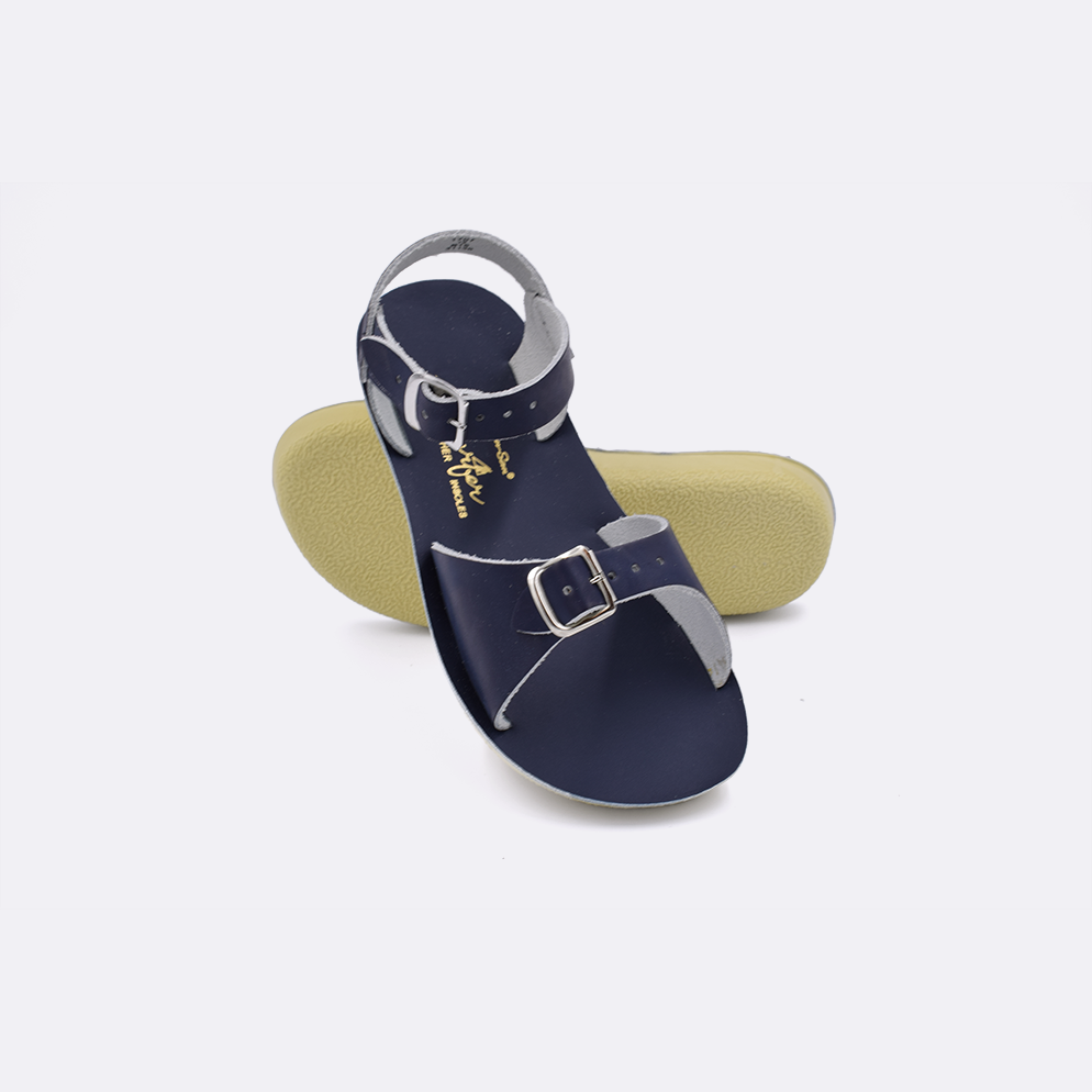 Two little kid sized 1700 Surfer style sandals with navy straps and navy insoles.  One standing with the sole facing the camera. The second is laying diagonally over the top left edge of the sole.