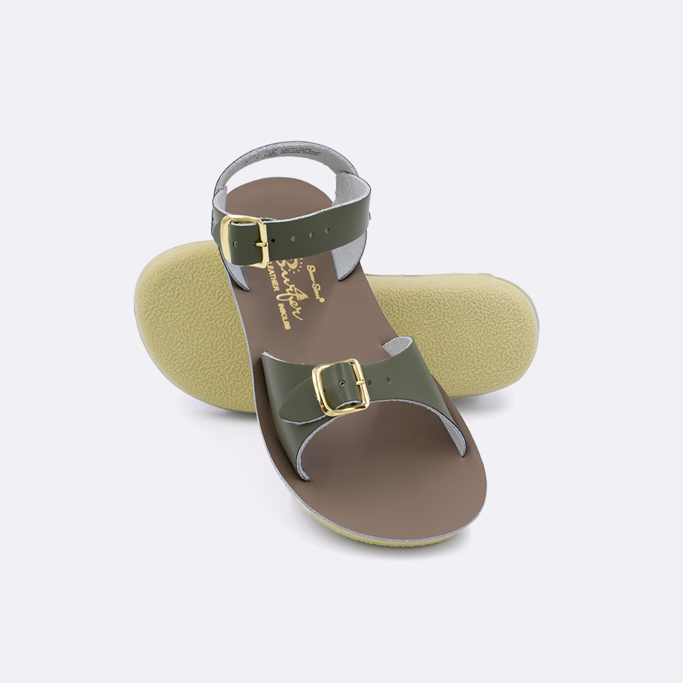 Two little kid sized 1700 Surfer style sandals with olive straps and beige insoles.  One standing with the sole facing the camera. The second is laying diagonally over the top left edge of the sole.