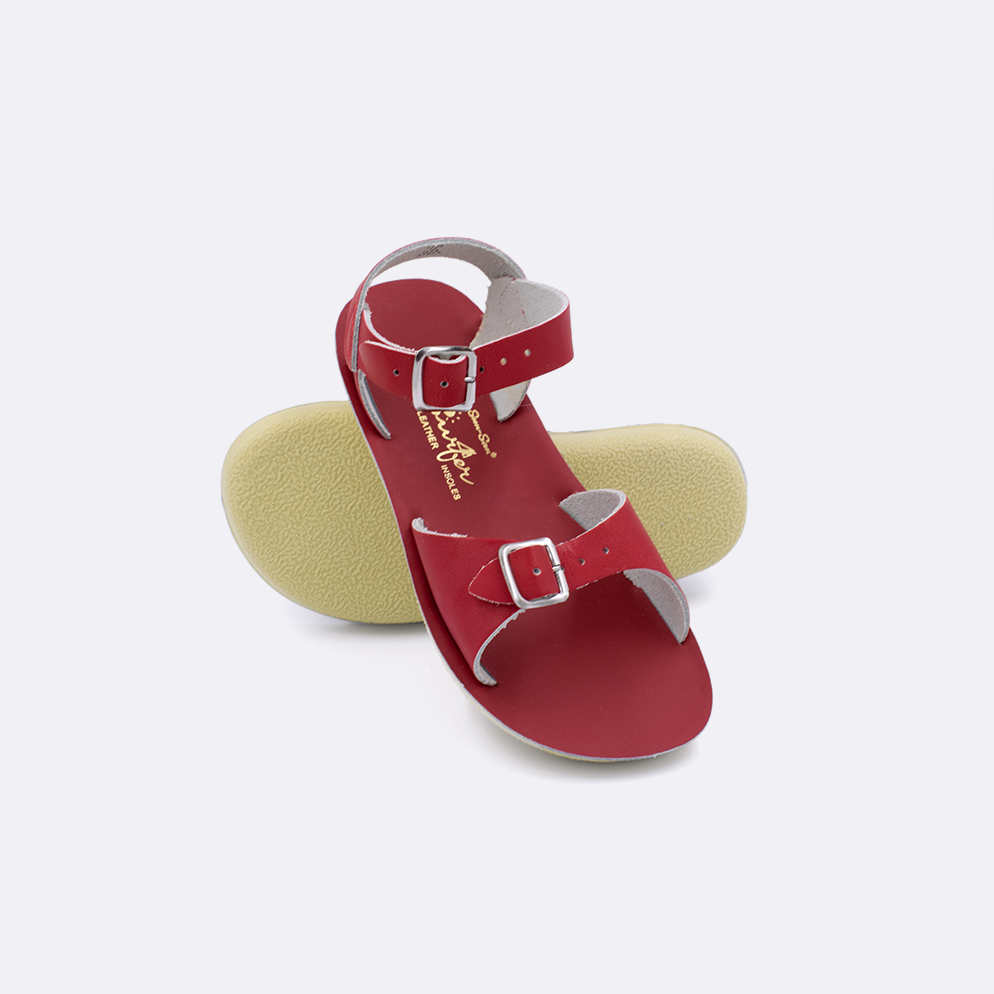 Two little kid sized 1700 Surfer style sandals with red straps and red insoles.  One standing with the sole facing the camera. The second is laying diagonally over the top left edge of the sole.