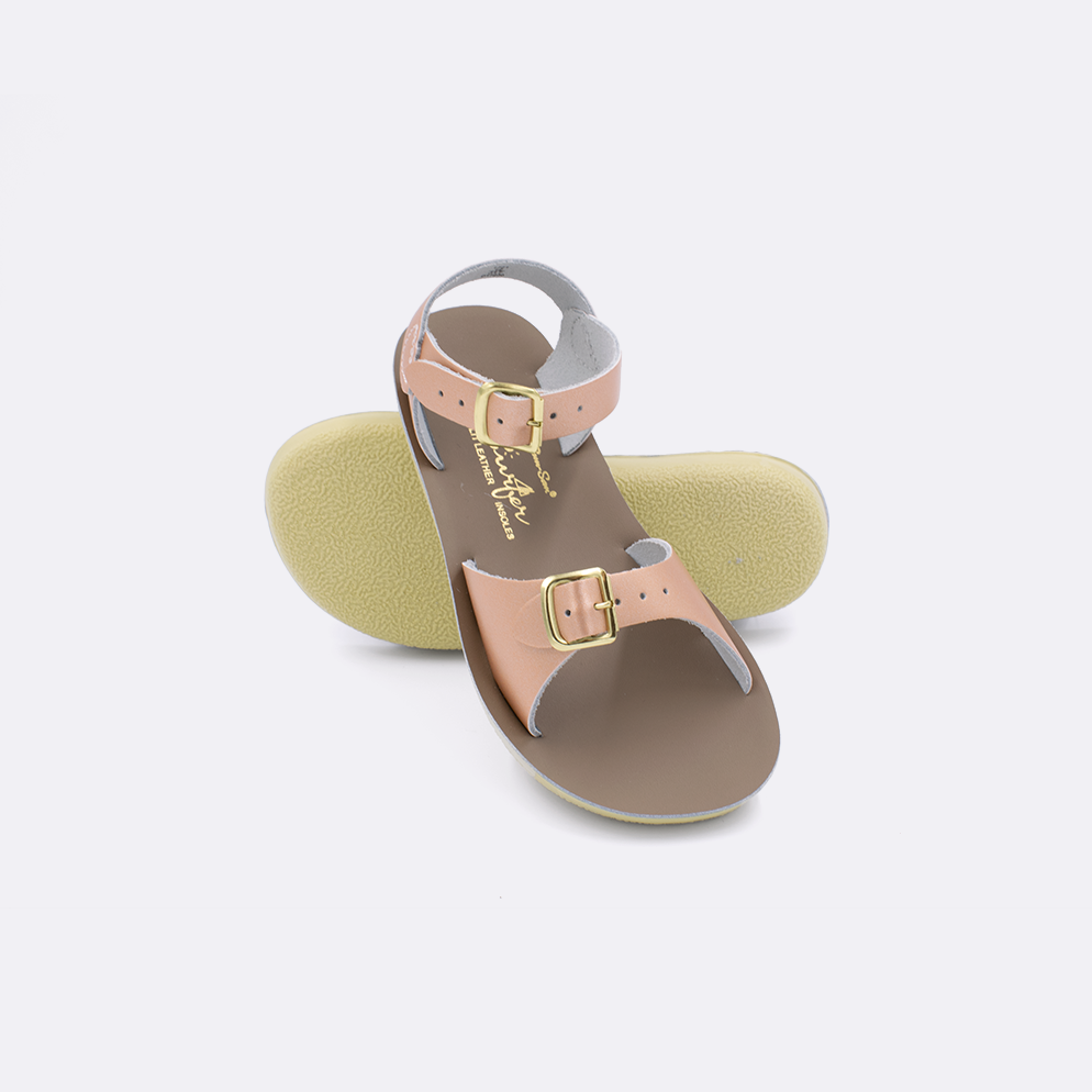 Two little kid sized 1700 Surfer style sandals with rose gold straps and beige insoles.  One standing with the sole facing the camera. The second is laying diagonally over the top left edge of the sole.