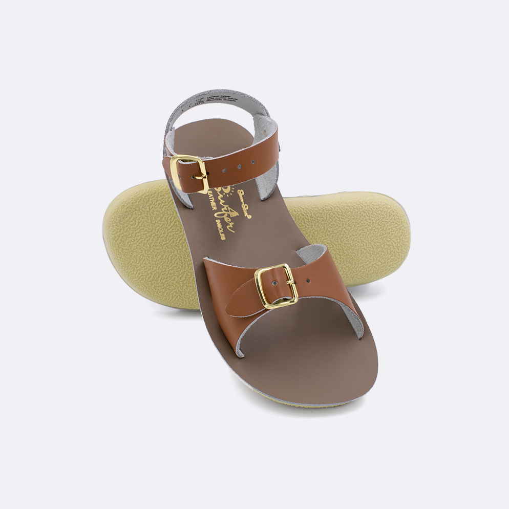 Two little kid sized 1700 Surfer style sandals with tan straps and beige insoles.  One standing with the sole facing the camera. The second is laying diagonally over the top left edge of the sole.