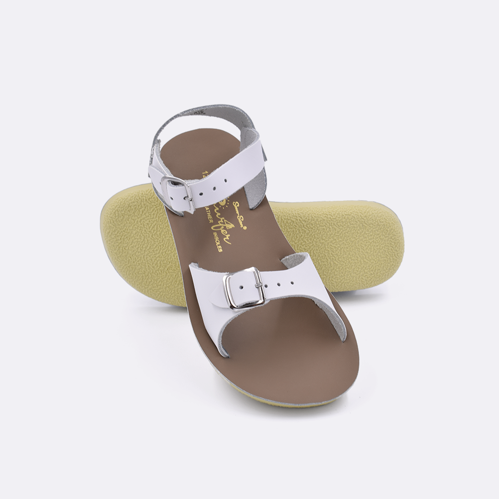 Two little kid sized 1700 Surfer style sandals with white straps and beige insoles.  One standing with the sole facing the camera. The second is laying diagonally over the top left edge of the sole.