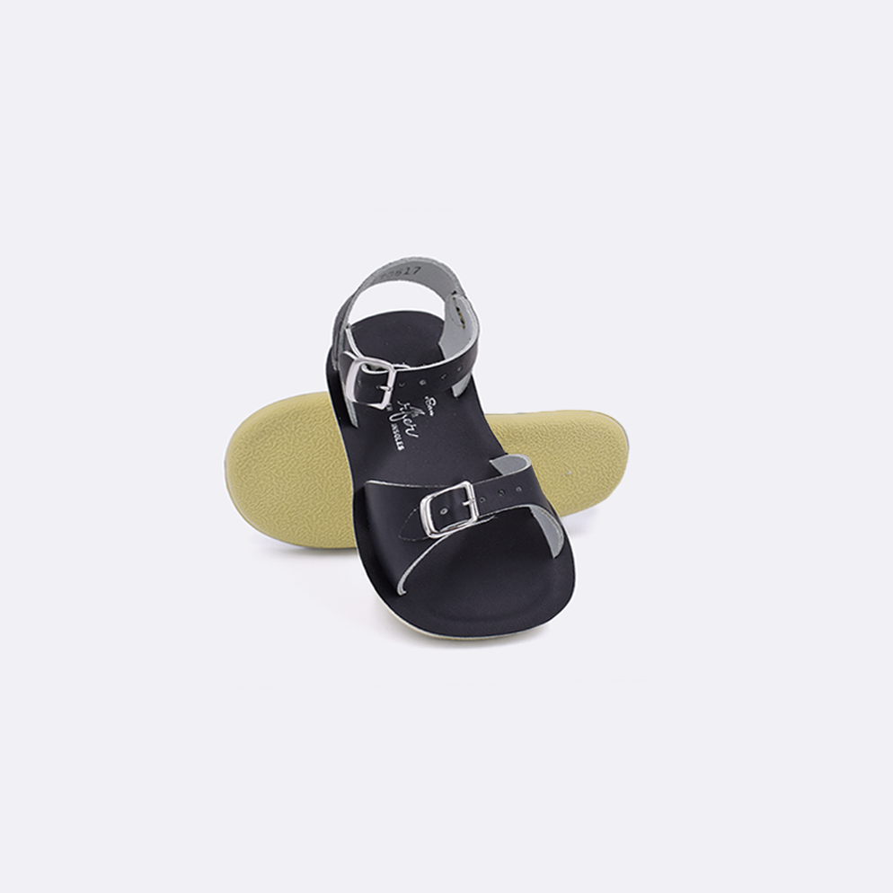 Two toddler sized 1700 Surfer style sandals with black straps and black insoles.  One standing with the sole facing the camera. The second is laying diagonally over the top left edge of the sole.