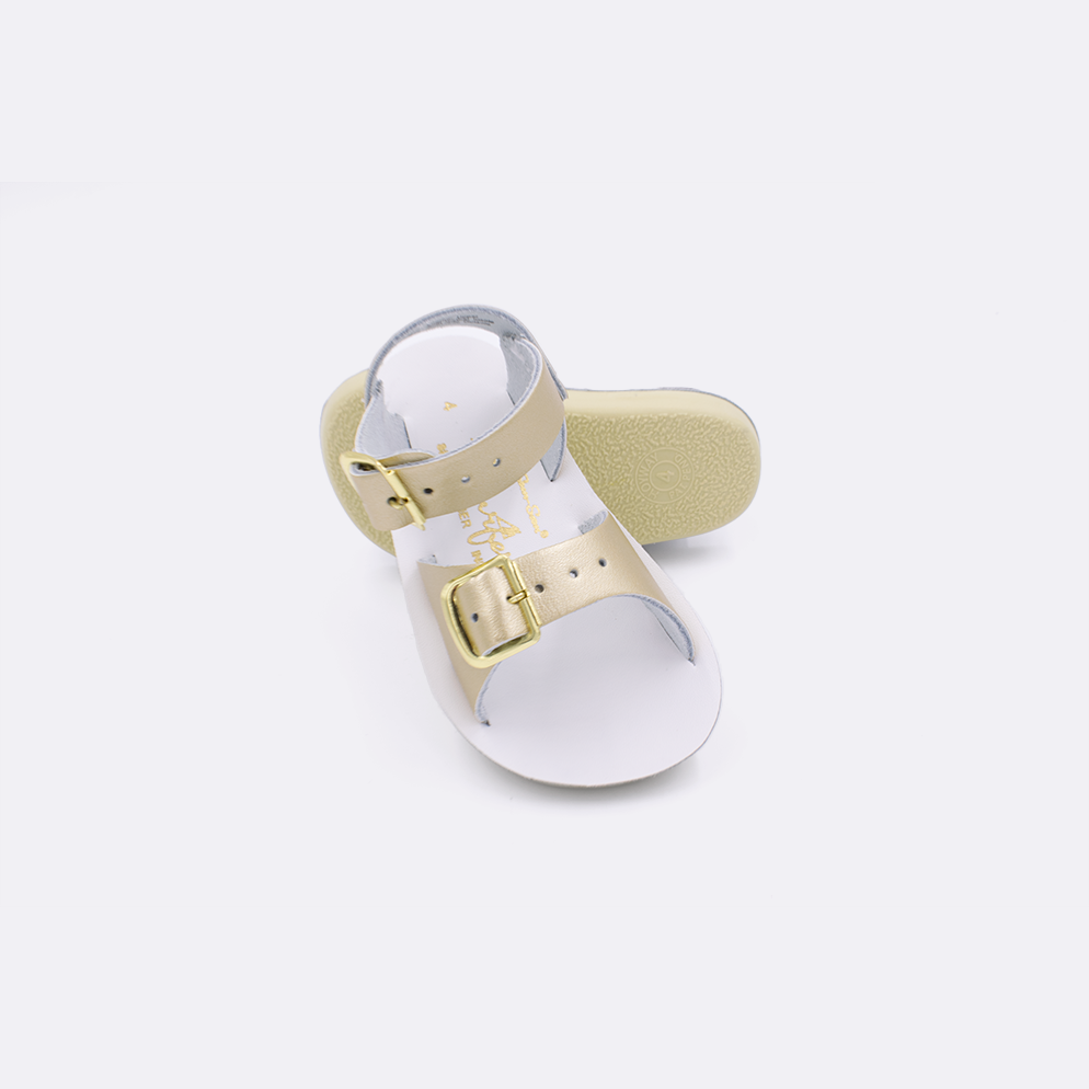 Two baby sized 1700 Surfer style sandals with gold straps and white insoles.  One standing with the sole facing the camera. The second is laying diagonally over the top left edge of the sole.