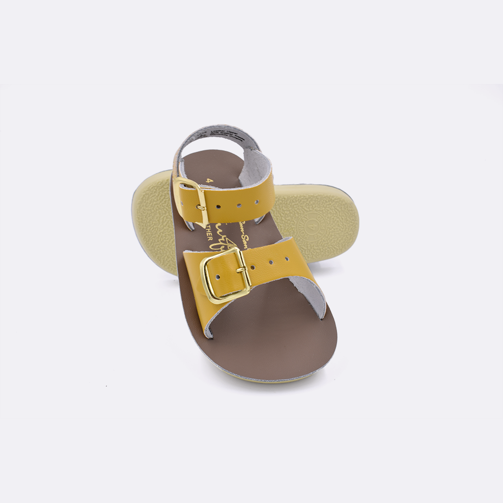 Two baby sized 1700 Surfer style sandals with mustard straps and beige insoles.  One standing with the sole facing the camera. The second is laying diagonally over the top left edge of the sole.