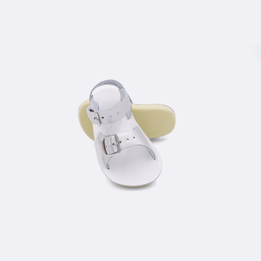 Two baby sized 1700 Surfer style sandals with silver straps and white insoles.  One standing with the sole facing the camera. The second is laying diagonally over the top left edge of the sole.
