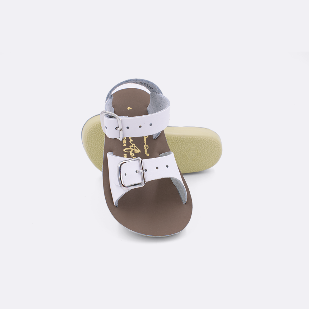 Two baby sized 1700 Surfer style sandals with white straps and beige insoles.  One standing with the sole facing the camera. The second is laying diagonally over the top left edge of the sole.