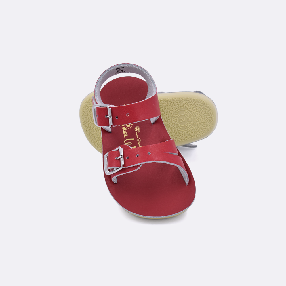 Two baby sized 2000 Sea Wee style sandals with red straps and red insoles.  One standing with the sole facing the camera. The second is laying diagonally over the top left edge of the sole.