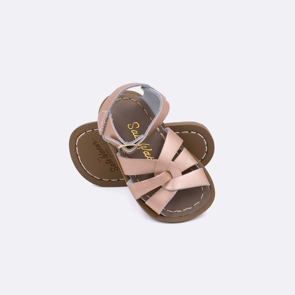 Two baby size 800 Original style sandals color rose gold.  One standing with the sole facing the camera. The second is laying diagonally over the top left edge of the sole.