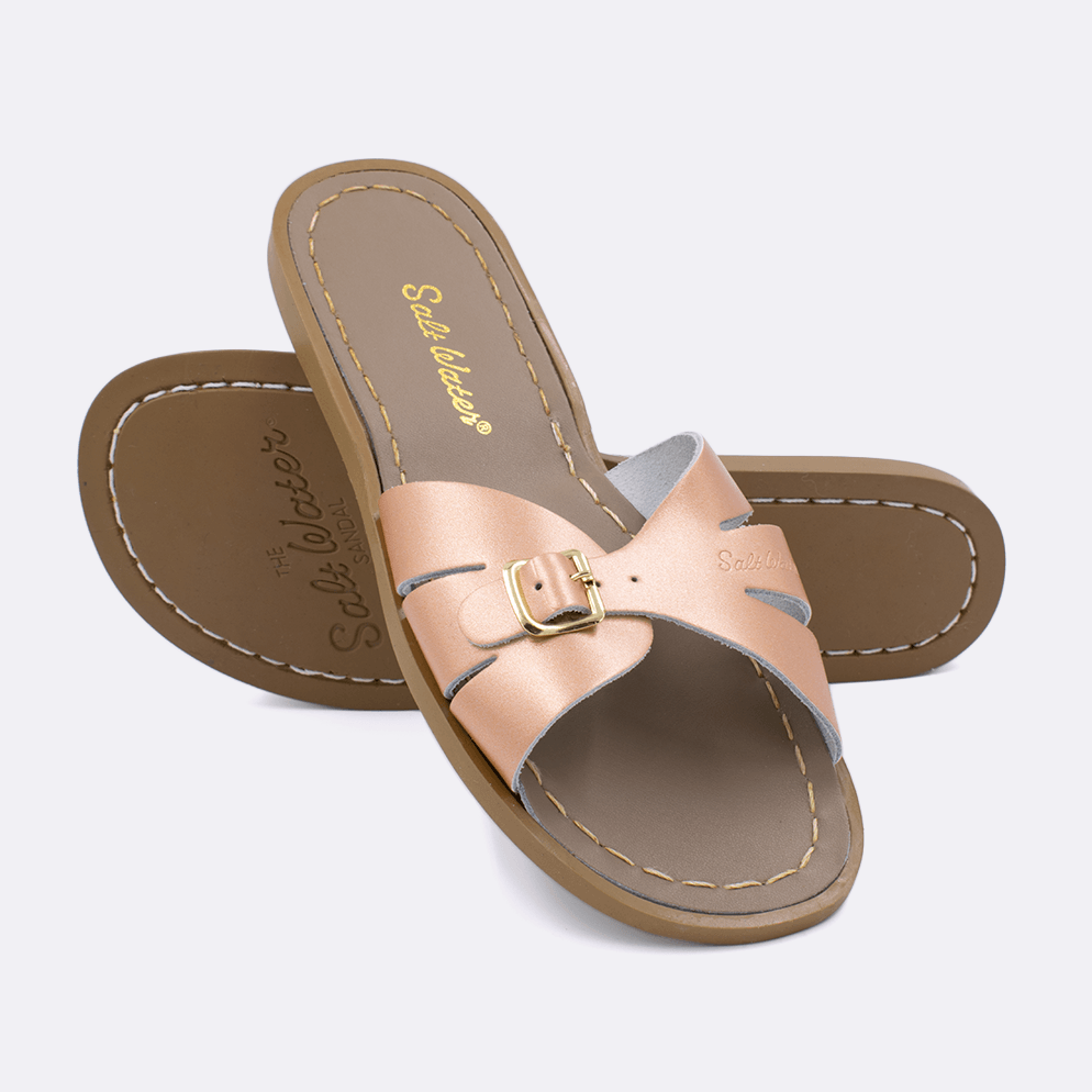 Two 9900 Classic Slide style sandals color rose gold.  One standing with the sole facing the camera. The second is laying diagonally over the top left edge of the sole.	Adult Size.