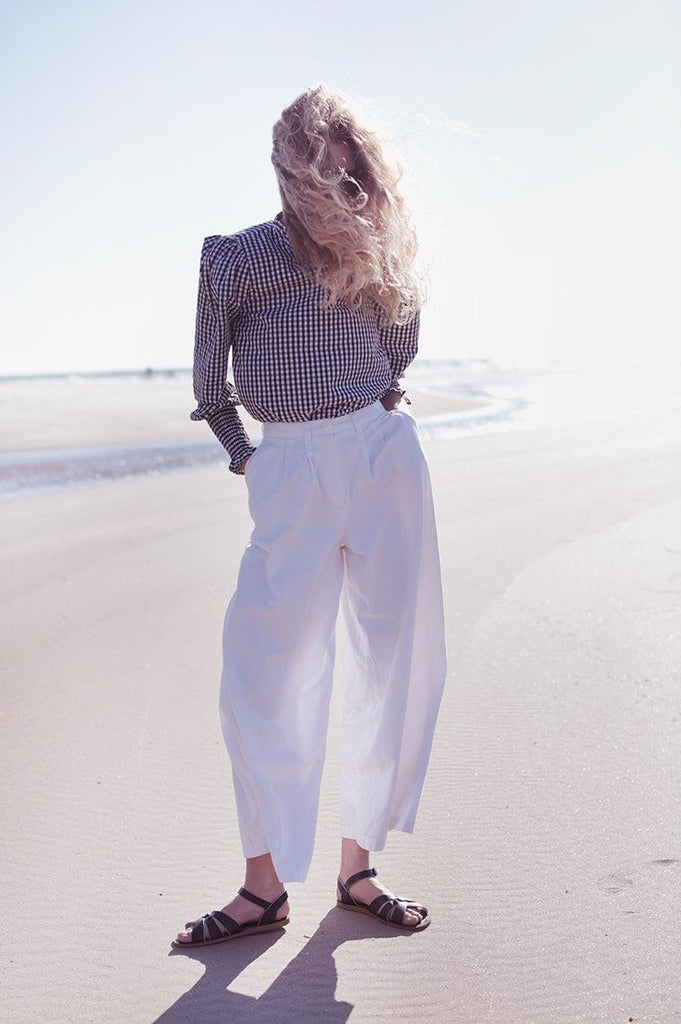 An adult with blonde hair is standing on the beach with a checkered black and white shirt tucked into white pants. Their hands are in their pockets with their feet diagonally facing each direction wearing our Brown Salt Water Originals.