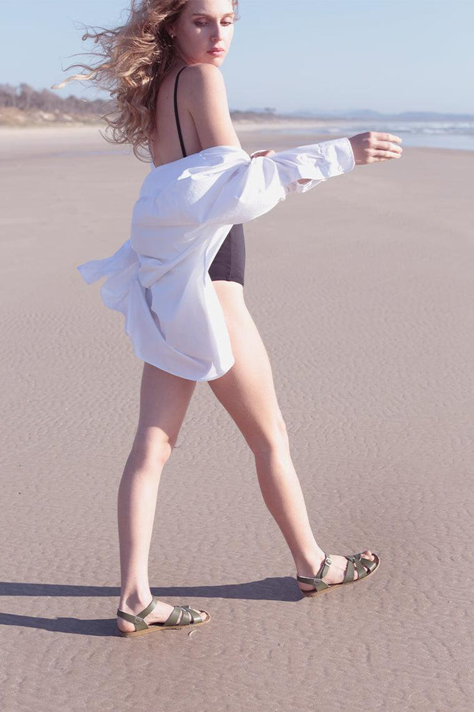 An adult with blonde long blond hair wearing a black one piece swimsuit and a white coverup. They are walking on the beach wearing our olive salt water originals.