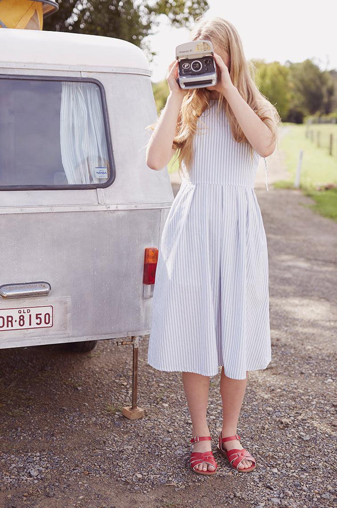 An adult with long blonde hair is wearing a light blue and white striped dress. They are taking a picture with a camera while standing in front of an old wahite and grey van. they are wearing our red salt water original sandals.