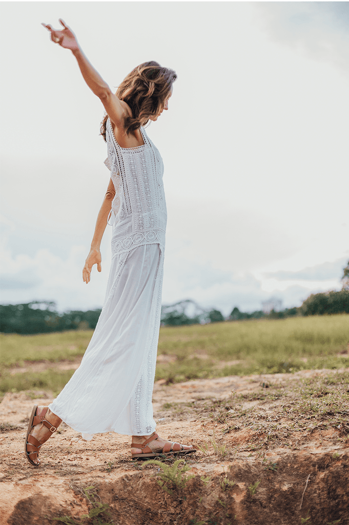 An adult with long brown hair walking on a dirt and grass terrain. They are wearing a white dress and our tan salt water original sandals while swinging their right arm in the air. 