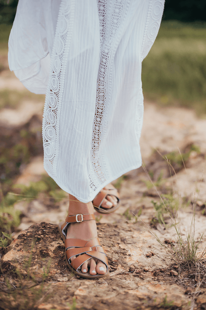 A Close up photo of an adults legs wearing a white dress and our tan salt water originals with the right foot in front of the left. They are walking on a dirt and grassy terrain. 