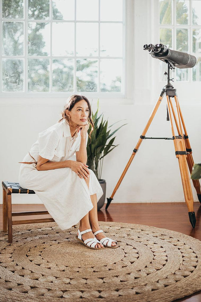 An adult with long brown is wearing a white button down dress.  They are wearing our white salt water original sandals. The room has two windows, a white wall, brown hardwood floors, and a woven rug.